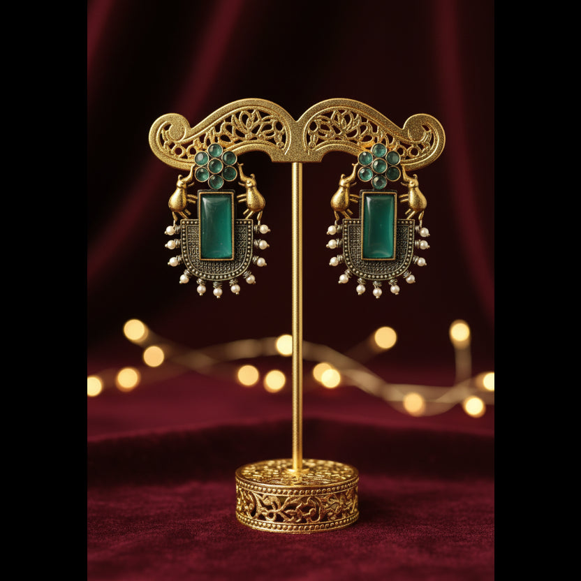 A pair of ornate gold-toned earrings with rectangular green turquoise-like stones and small bead details, displayed on a textured gray fabric background.