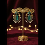 A pair of ornate gold-toned earrings with rectangular green turquoise-like stones and small bead details, displayed on a textured gray fabric background.