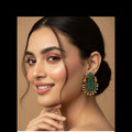 A close-up of a woman wearing a large ornate gold-toned statement earring with a green rectangular stone and small pearl-like accents, against a warm beige studio background.