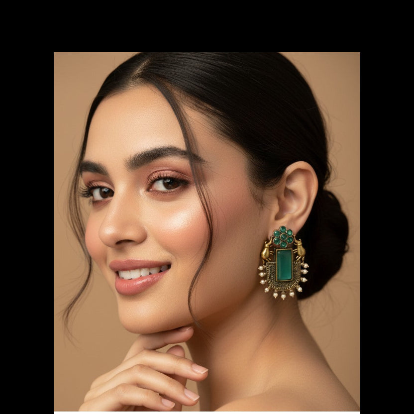 A close-up of a woman wearing a large ornate gold-toned statement earring with a green rectangular stone and small pearl-like accents, against a warm beige studio background.