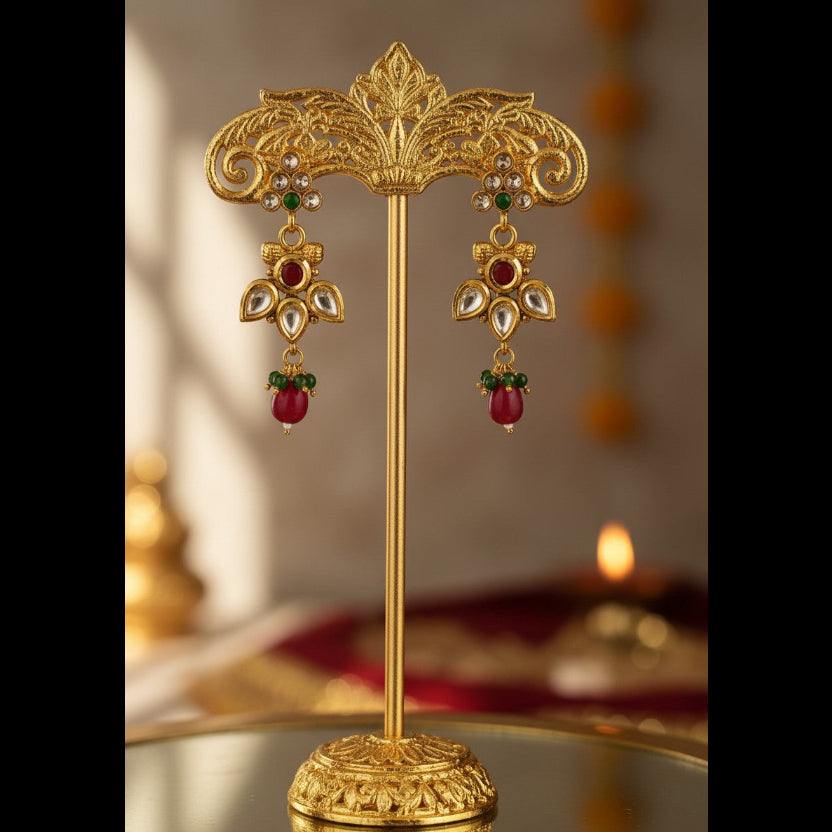 An ornate pair of gold-toned traditional earrings displayed on a matching gold stand. The earrings feature red bead drops, green stone accents, and clear stone details, set against a softly blurred warm background.