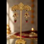 An ornate pair of gold-toned traditional earrings displayed on a matching gold stand. The earrings feature red bead drops, green stone accents, and clear stone details, set against a softly blurred warm background.