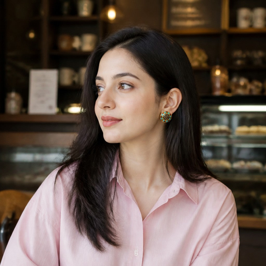 A woman with long dark hair sits at a wooden table in a cafe, wearing a light pink button-down shirt and blue jeans. A white coffee cup is on the table in the foreground, and shelves with coffee equipment are visible in the background.