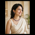 A woman with dark hair smiles while wearing a cream-colored draped top and a colorful beaded necklace with matching square earrings. The background is a softly blurred indoor setting with warm beige tones.