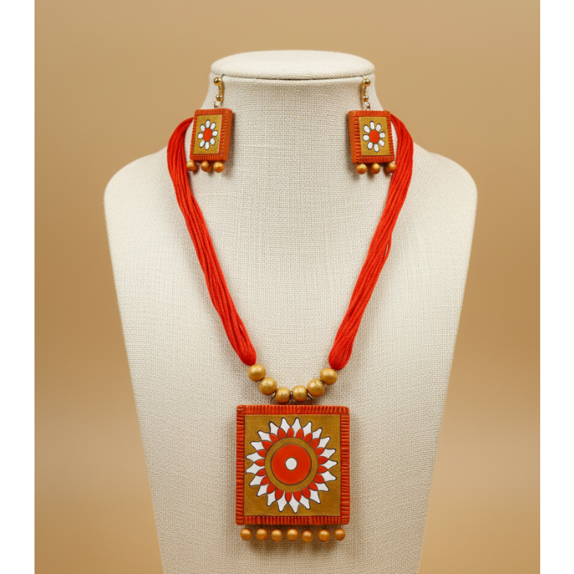 A decorative jewelry set featuring square gold-toned pendants and matching earrings with hand-painted floral motifs in orange, white, and red, accented with red tassels and gold beads, shown on a dark textured background.