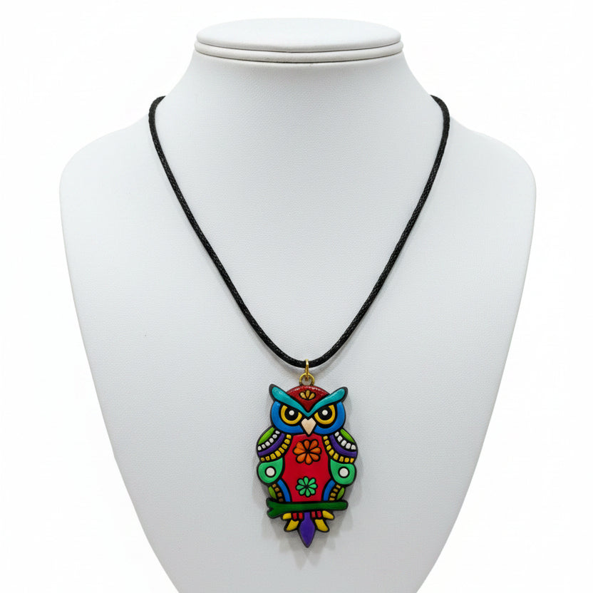 A colorful owl pendant necklace on a black cord, displayed on a white card in a clear plastic sleeve, surrounded by other jewelry items on a blue patterned surface.