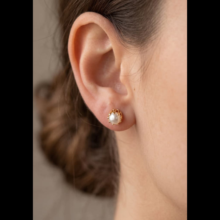 A close-up of a woman's ear wearing a small gold stud earring with a white pearl-like center, against a soft neutral background.