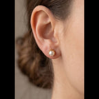 A close-up of a woman's ear wearing a small gold stud earring with a white pearl-like center, against a soft neutral background.