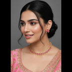 A woman with dark hair styled in a low bun wears a gold-toned necklace and matching flower-shaped earrings with pink stones, paired with a pink embroidered top against a plain gray studio background.