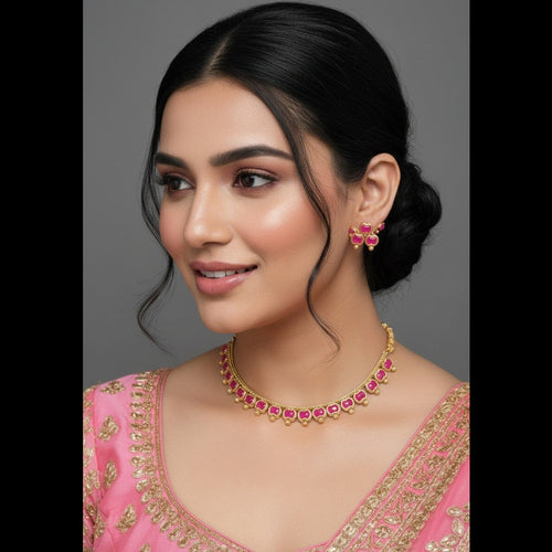 A woman with dark hair styled in a low bun wears a gold-toned necklace and matching flower-shaped earrings with pink stones, paired with a pink embroidered top against a plain gray studio background.