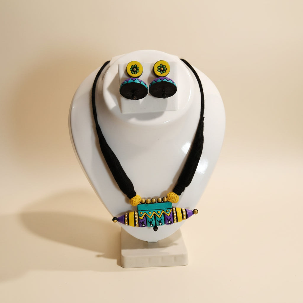 A handmade jewelry set featuring a beaded necklace and matching earrings displayed on a white ceramic jewelry stand. The jewelry features a black cord necklace with a colorful beaded bar pendant and matching stud earrings with yellow, purple, green, and black patterns.