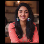 A smiling woman with long dark hair wearing colorful, ornate jhumka earrings and a coral pink embroidered top, photographed in a softly blurred indoor setting.