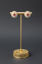A pair of round stud earrings with a gold-toned halo of small clear stones and a central red stone, displayed on a white block stand with a black vertical post.