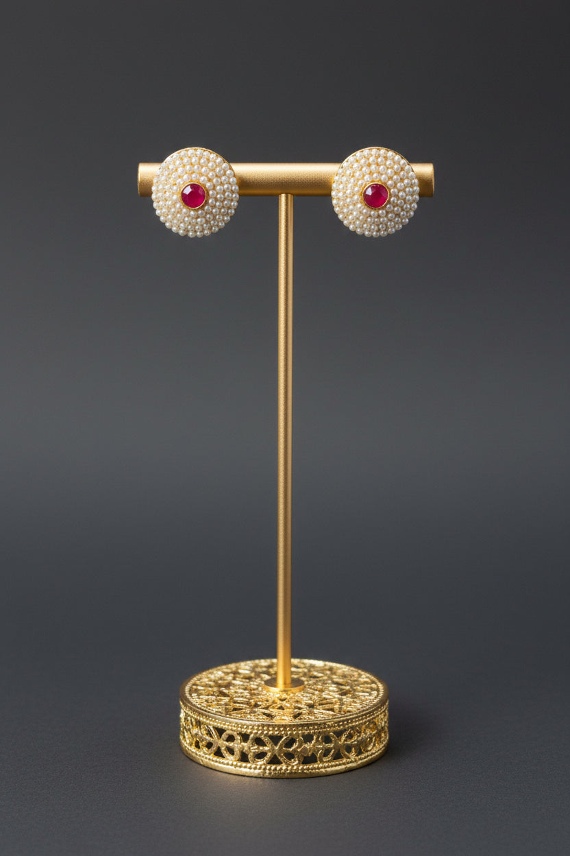 A pair of round stud earrings with a gold-toned halo of small clear stones and a central red stone, displayed on a white block stand with a black vertical post.