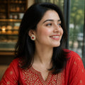 A woman wearing a red embroidered traditional outfit with gold detailing, seated indoors near a window. She is smiling and looking to the side, with her hair styled in loose waves and a red-and-gold earring visible.