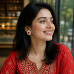 A woman wearing a red embroidered traditional outfit with gold detailing, seated indoors near a window. She is smiling and looking to the side, with her hair styled in loose waves and a red-and-gold earring visible.